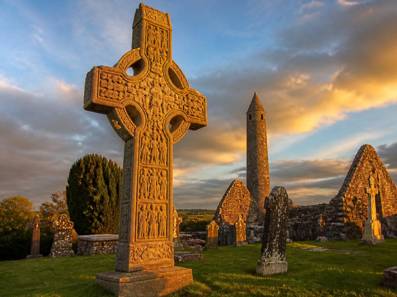 A tall ringed Irish high cross carved with biblical scenes standing in a monastic graveyard at golden hour, with a round tower behind